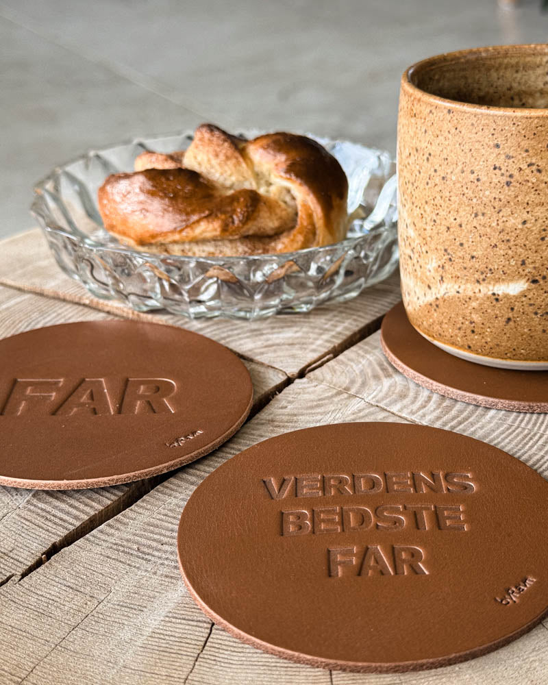 MOKKA Untersetzer aus cognacfarbenem Leder mit Prägung "Far" und "Verdens bedste far" neben Kaffeetasse und Zimtschnecke auf Holztisch