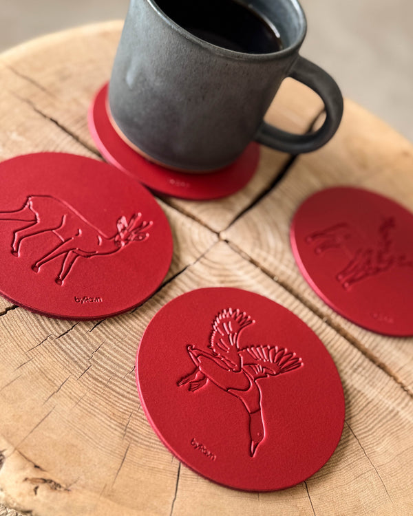 MOKKA Jagd-Untersetzer aus rotem Leder mit geprägter fliegender Stockente, Rehbock und Hase auf Holzscheibe neben grauer Kaffeetasse
