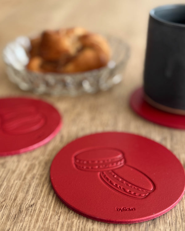 MOKKA Kaffee & Kuchen-Untersetzer aus rotem Leder mit geprägten Macarons auf Holztisch neben schwarzer Tasse und Zimtschnecke in Glasschale