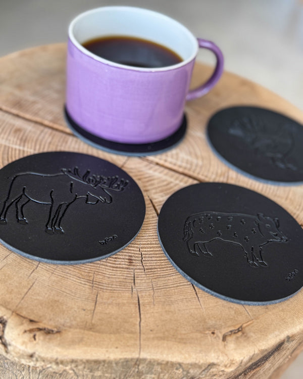 MOKKA Jagd-Untersetzer aus schwarzem Leder mit geprägtem Elch, Wildschwein und Auerhahn auf Holzscheibe mit lila Kaffeetasse auf Untersetzer