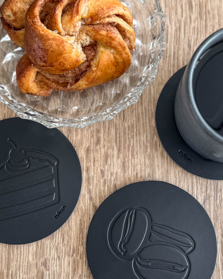 MOKKA Kaffee & Kuchen-Untersetzer aus schwarzem Leder mit geprägter Torte und Kaffeebohnen auf Holztisch neben Zimtschnecke und grauer Tasse