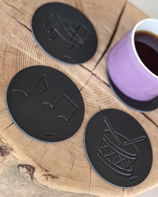 MOKKA Untersetzer aus schwarzem Leder, geprägt mit Noten, Trommel und Flügel, auf Holzscheibe mit lila Kaffeetasse