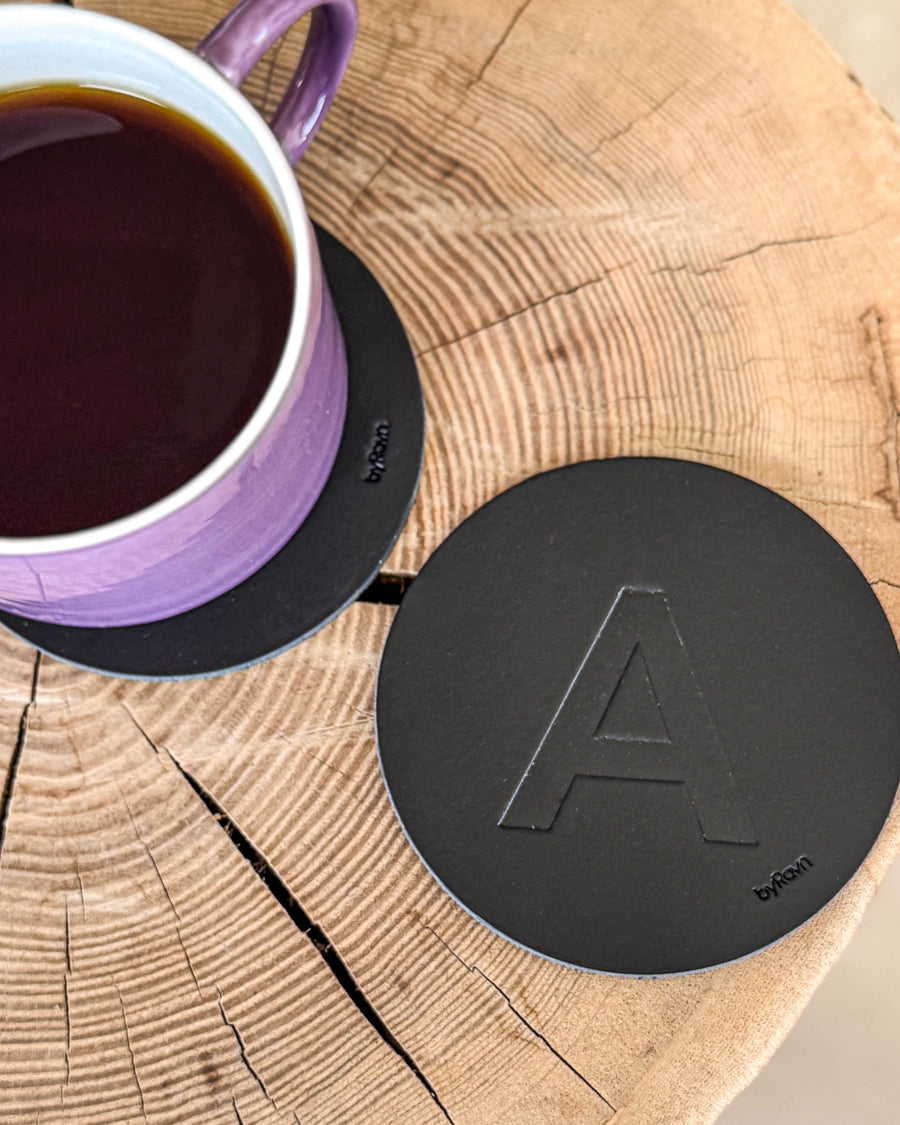 MOKKA Untersetzer aus schwarzem Leder mit geprägtem Buchstaben "A" auf Holzscheibe neben lila Kaffeetasse auf Untersetzer