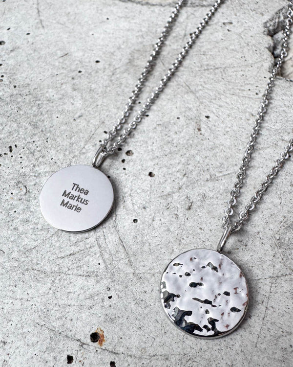 MOON QUOTE Namenskette aus silberfarbenem Edelstahl mit den Namen "Thea Markus Marie" graviert, gehämmerte Seite daneben, auf Beton