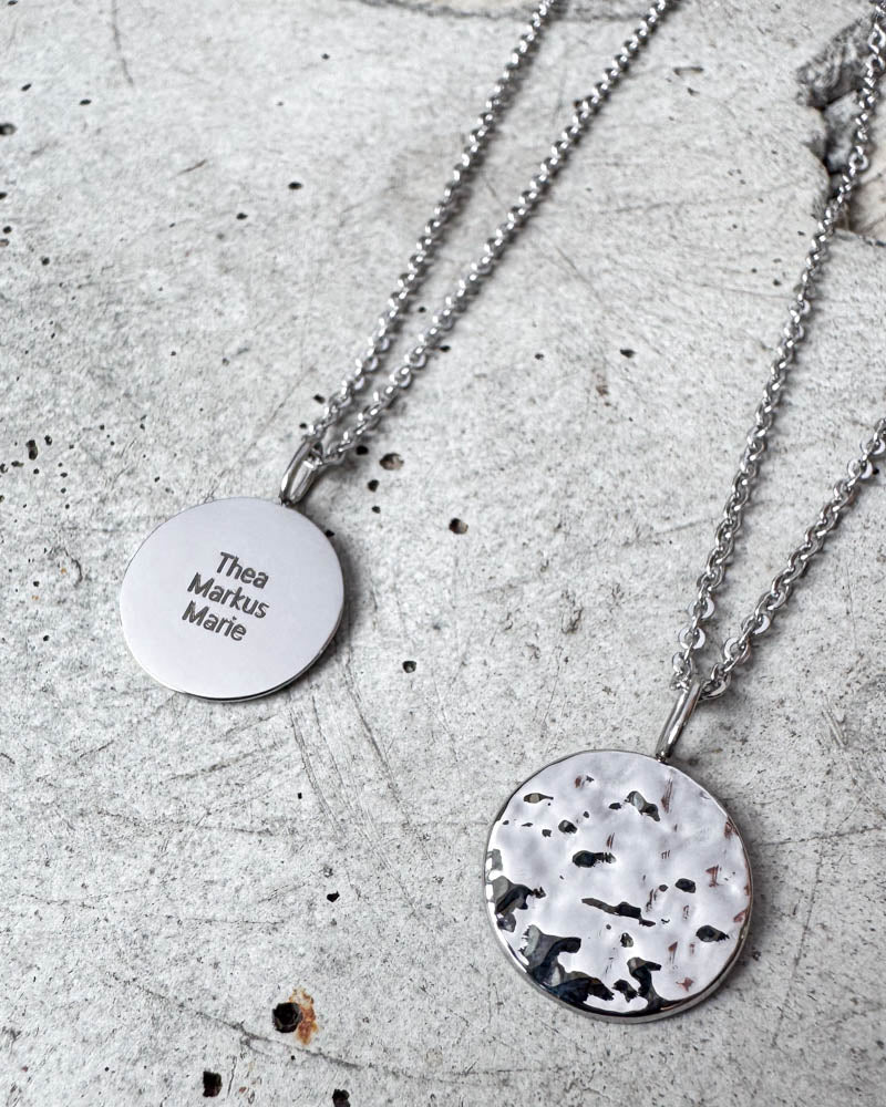 MOON QUOTE Namenskette aus silberfarbenem Edelstahl mit den Namen "Thea Markus Marie" graviert, gehämmerte Seite daneben, auf Beton
