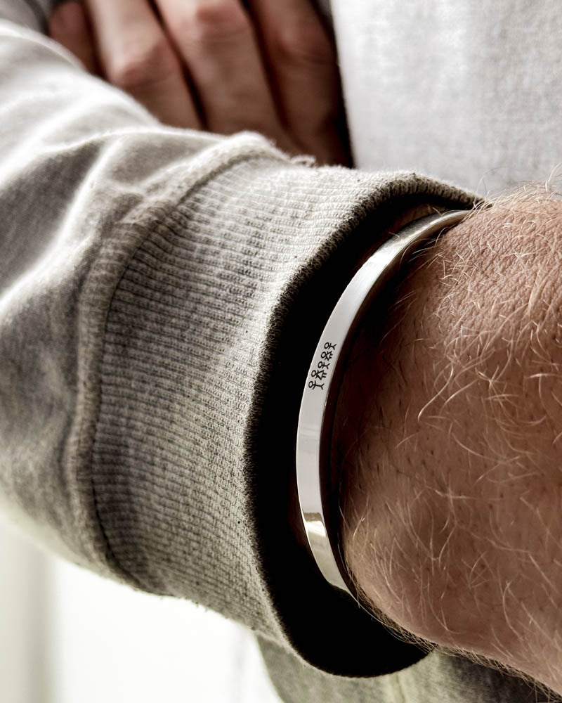 MARS STICK Armband aus silberfarbenem Edelstahl mit gravierter Streichholz-Familie am Handgelenk eines Mannes, hellgraues Sweatshirt