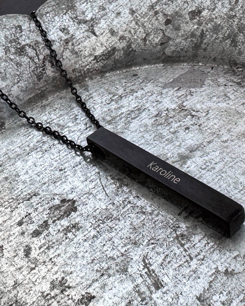 NEO Namenskette aus schwarzem Edelstahl mit dem Namen "Karoline" auf länglichem Stab graviert, auf patinierter Zinkschale liegend