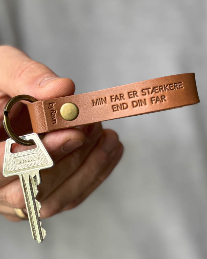 RAVN Schlüsselanhänger aus Cognac-Leder mit der Gravur "Min far er stærkere end din far", in der Hand gehalten mit Schlüssel