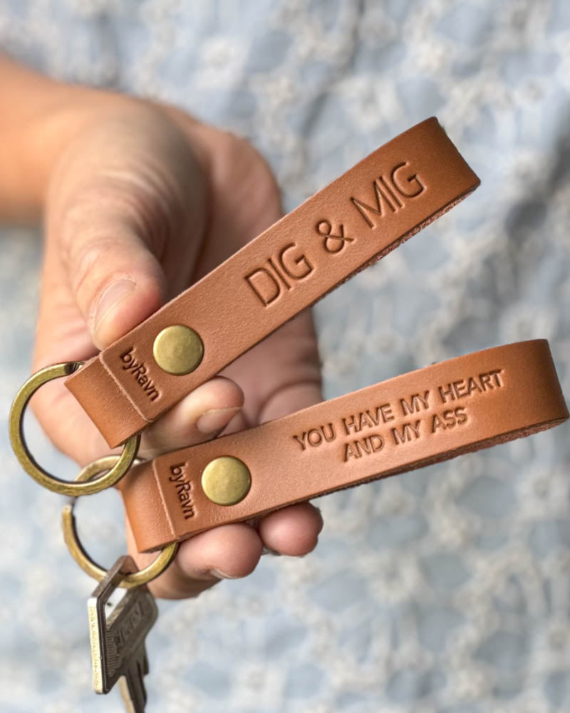 Zwei RAVN Schlüsselanhänger aus Cognac-Leder in der Hand gehalten mit den Gravuren "Dig & mig" und "You have my heart and my ass"