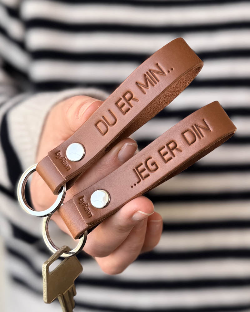 RAVN Schlüsselanhänger aus Cognac-Leder als Partnerset mit den Gravuren "Du er min.." und "..jeg er din", in der Hand gehalten mit Schlüssel