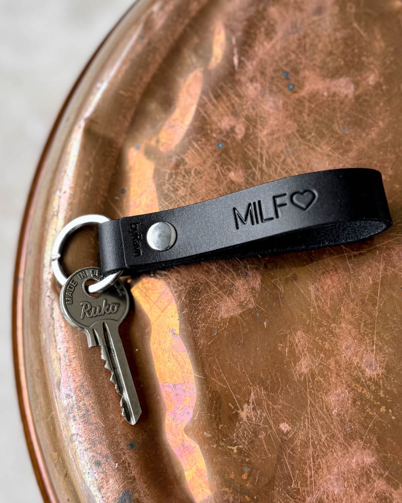 RAVN Schlüsselanhänger aus dunklem Leder mit der Gravur "MILF" ♡ und Schlüssel, liegend auf patiniertem Kupfertablett