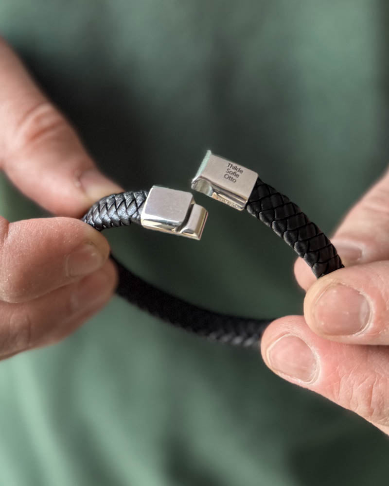 RAY Lederarmband aus schwarzem geflochtenem Leder mit offenem Magnetverschluss und den Namen "Thilde, Sofie, Otto" graviert, in den Händen gehalten