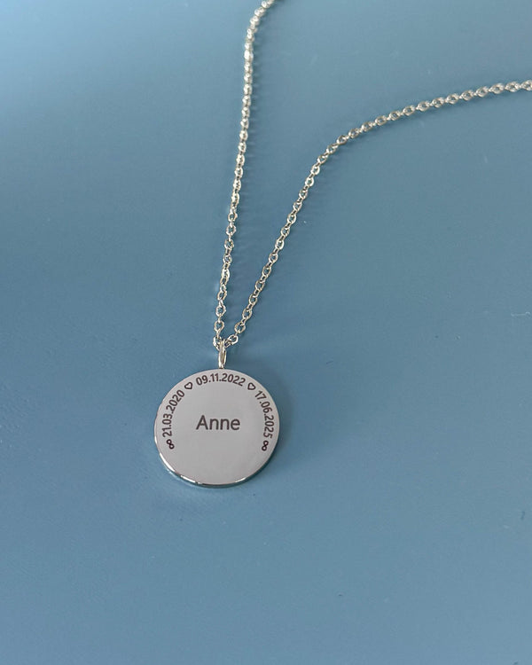 SIA CIRCLE Namenskette aus silberfarbenem Edelstahl mit "Anne" und den Daten "21.03.2020 ♡ 09.11.2022 ♡ 17.06.2025" im Kreis, auf blauem Grund