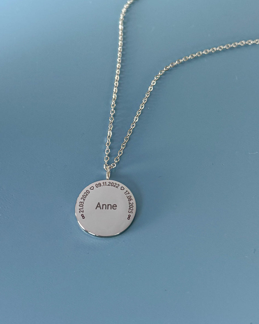 SIA CIRCLE Namenskette aus silberfarbenem Edelstahl mit "Anne" und den Daten "21.03.2020 ♡ 09.11.2022 ♡ 17.06.2025" im Kreis, auf blauem Grund