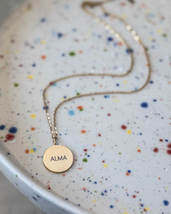 SIA NOTE Namenskette aus vergoldetem Edelstahl mit dem Namen "Alma" auf rundem Anhänger graviert, auf gesprenkelter Keramikschale