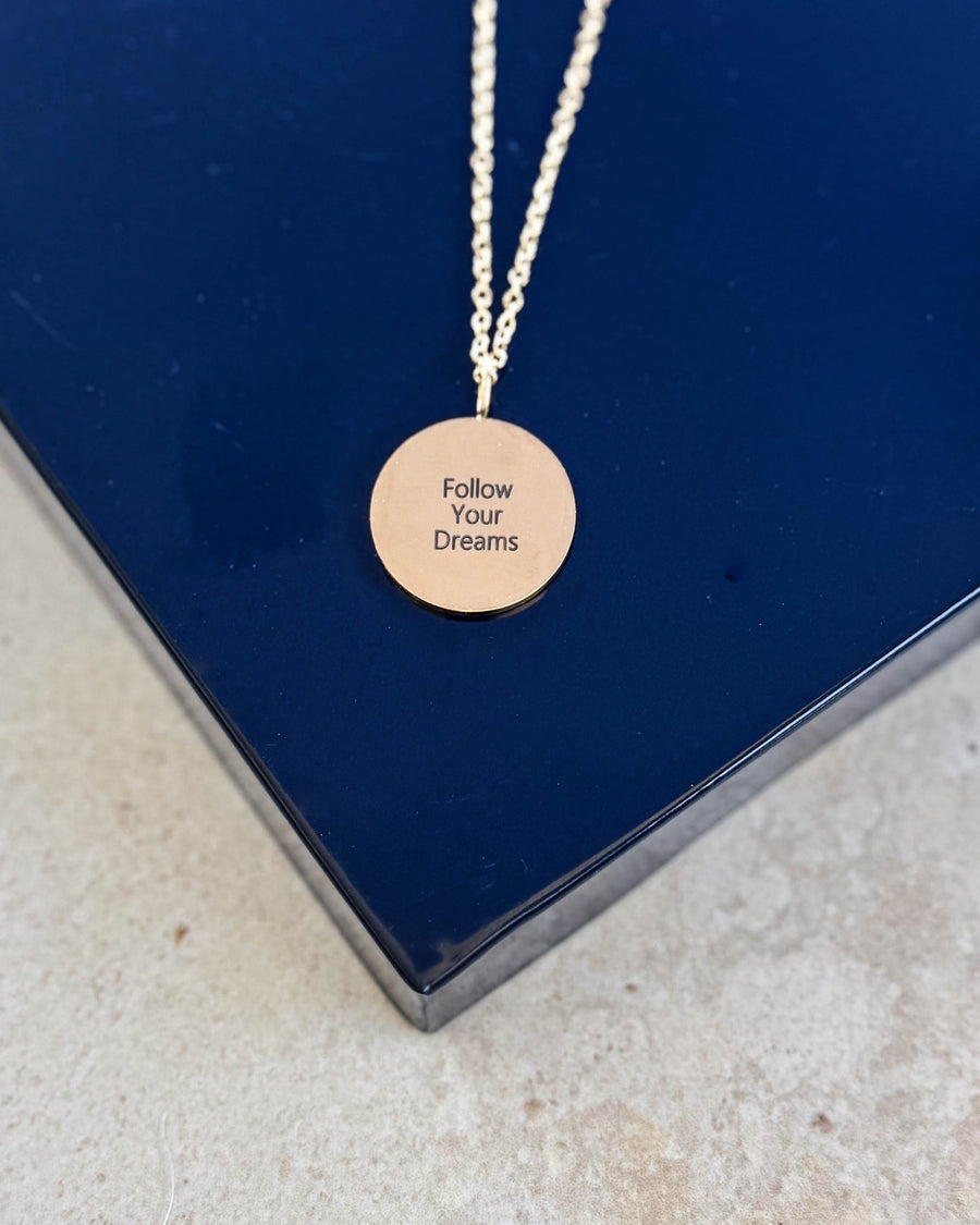 SIA QUOTE Halskette aus vergoldetem Edelstahl mit der Gravur "Follow your dreams" auf rundem Anhänger, auf dunkelblauer Schachtel