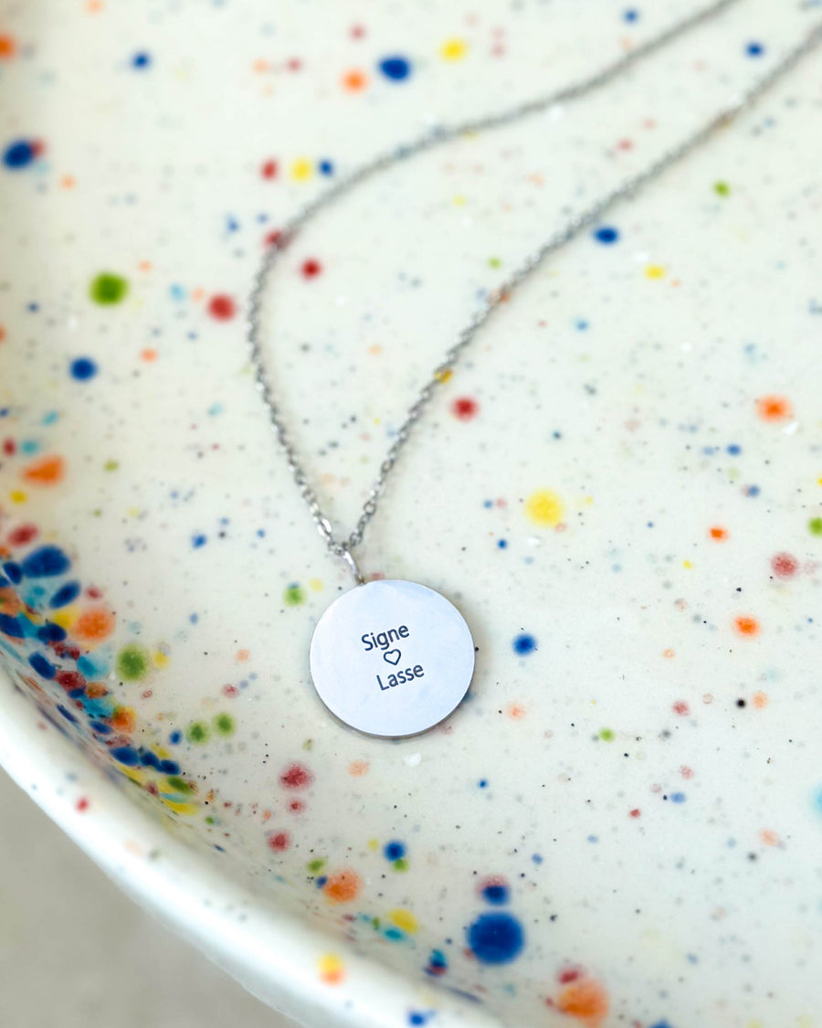 SIA QUOTE Namenskette aus silberfarbenem Edelstahl mit der Gravur "Signe ♡ Lasse" auf rundem Anhänger, auf gesprenkelter Schale