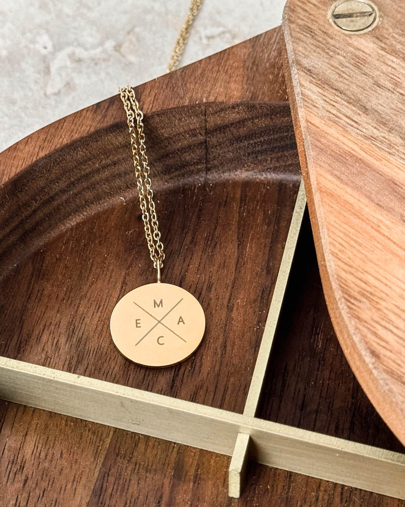 SIA X Halskette aus vergoldetem Edelstahl mit den Buchstaben "M E A C" kreuzförmig auf rundem Anhänger, in Holzschale mit Messingrand