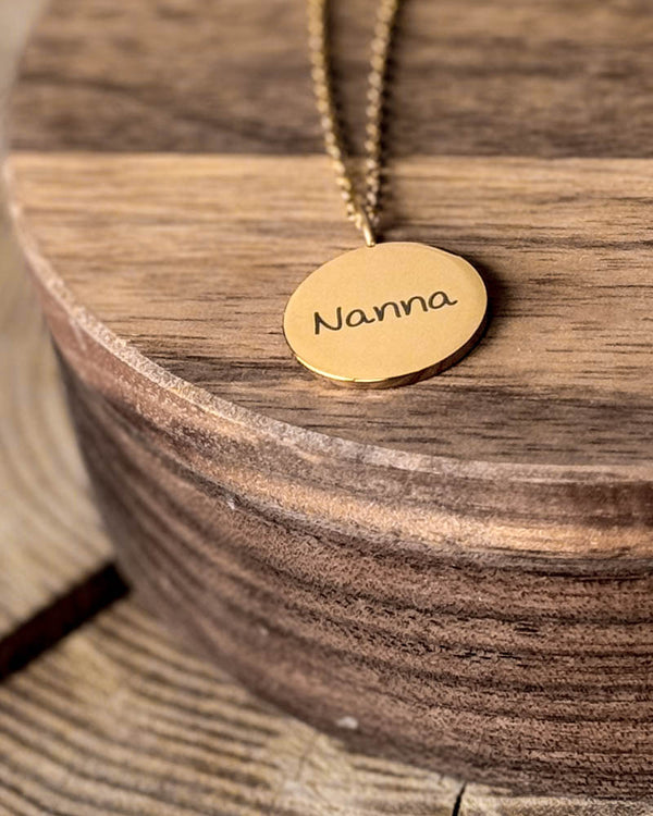 SIA NOTE SCRIPT Namenskette aus vergoldetem Edelstahl mit dem Namen "Nanna" in Handschrift auf rundem Anhänger graviert, auf runder Holzschale