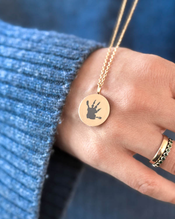 SIA MOTIF Halskette aus vergoldetem Edelstahl mit kleinem Handabdruck auf rundem Anhänger graviert, in der Hand gehalten über blauem Strick