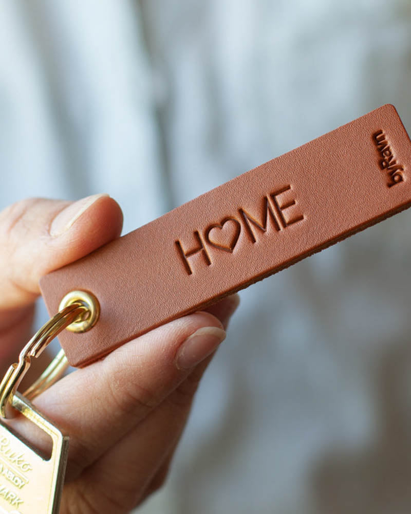SOLO Small Schlüsselanhänger aus Leder in Cognac, in der Hand gehalten, mit Schlüssel und der Gravur "Home", bei der das O ein Herz ist