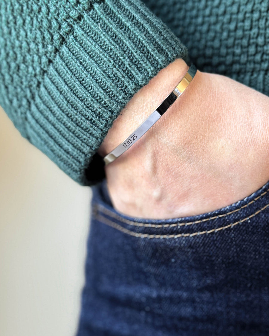 VENUS Armband aus silberfarbenem Edelstahl mit graviertem Datum "17.03.25", Hand in der Tasche dunkler Jeans, grüner Strickpullover