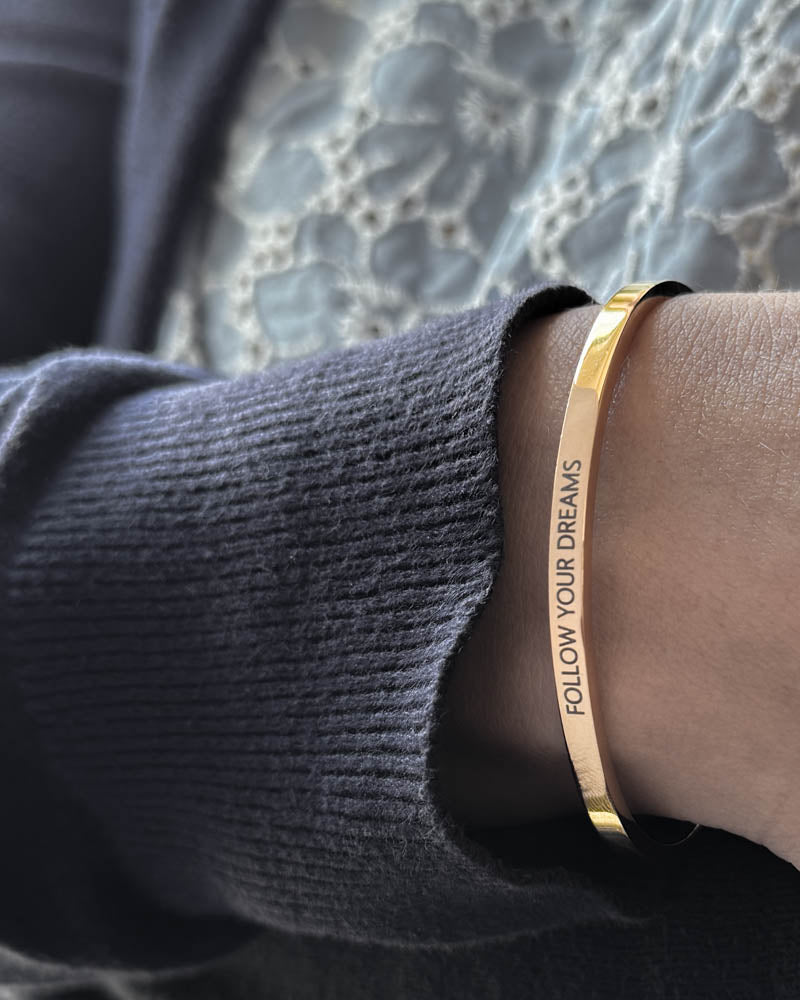 VENUS Armband aus vergoldetem Edelstahl mit der Gravur "Follow your dreams" in Großbuchstaben, zu dunkelblauem Strick und Spitzenbluse